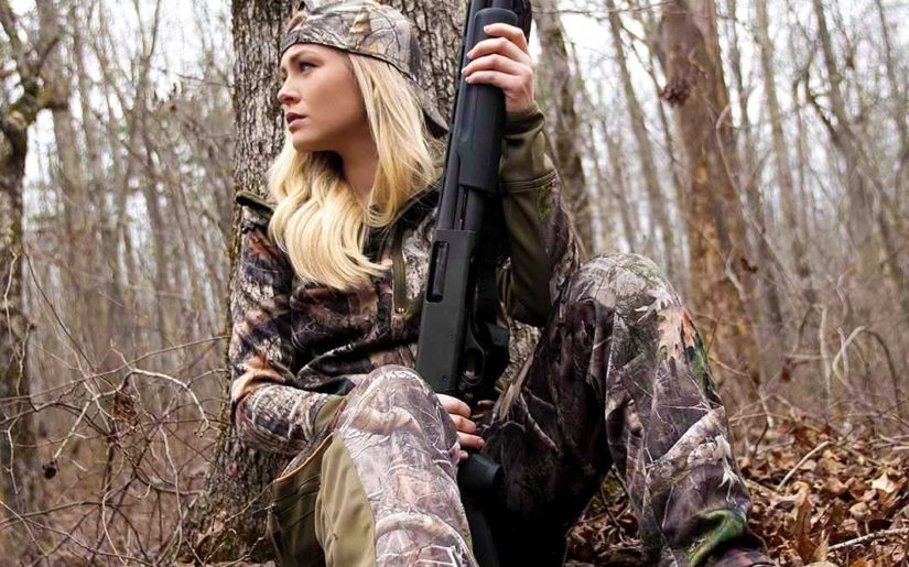 Campfire Collective Hunting Ambassador Ashley Dill wearing camouflage, holding a shotgun, leaning against a tree.
